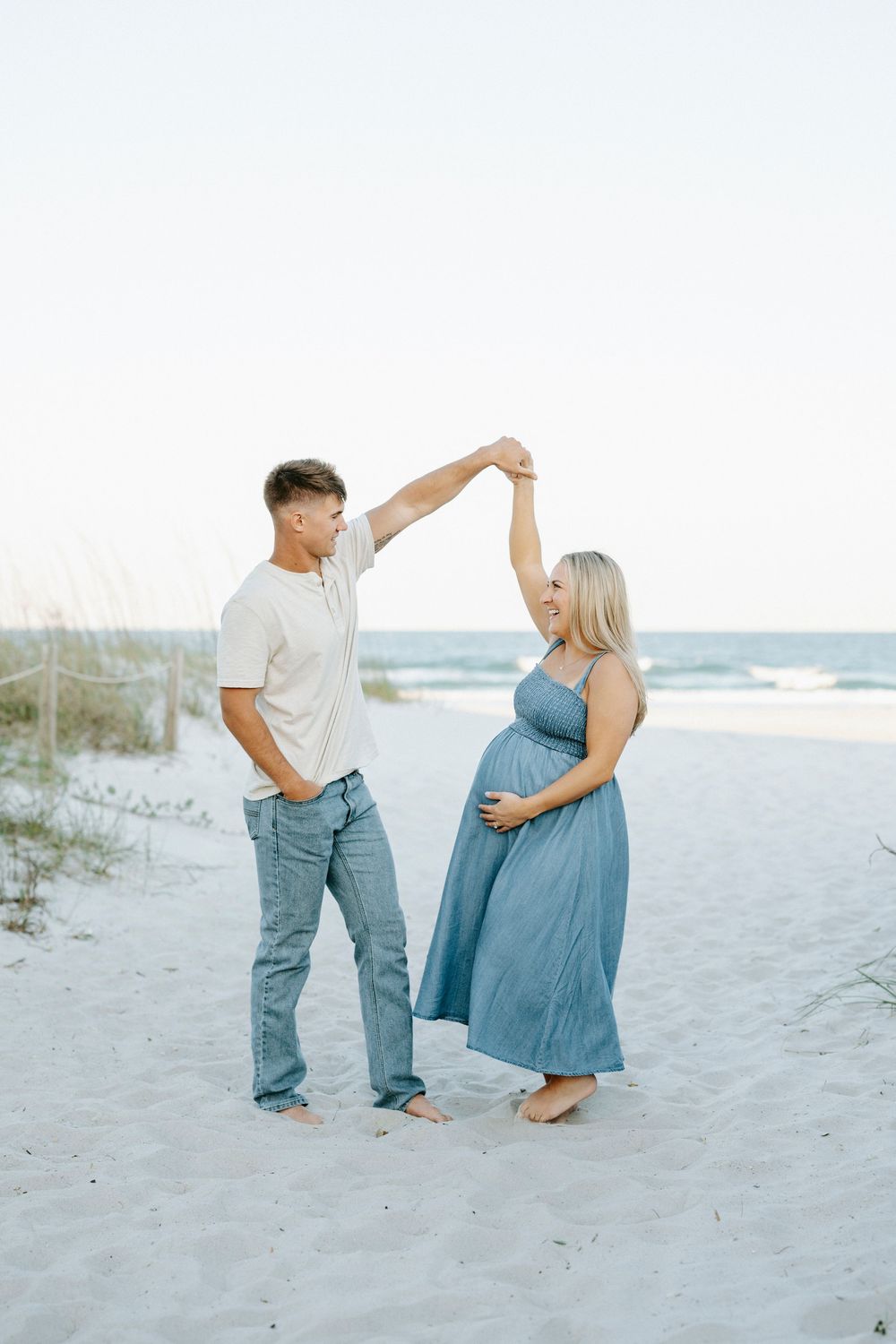 Pregnant couple dancing on a sandy beach, woman in blue maxi dress holding her belly, man in white shirt and jeans.