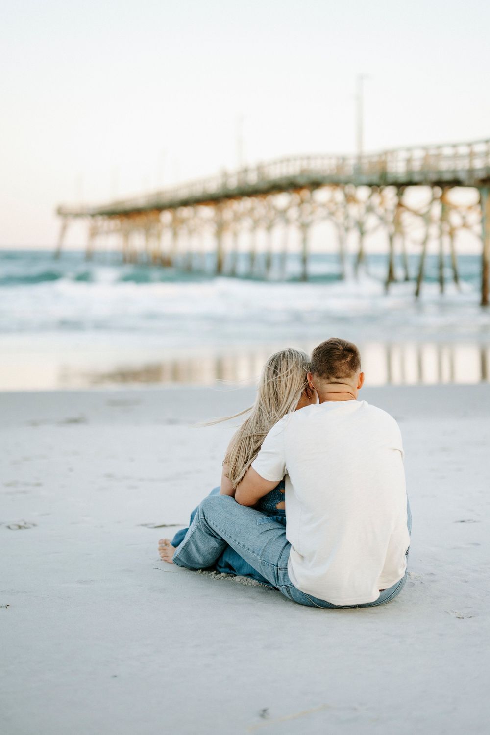 Couple sitting together on sandy beach near pier at sunset, woman leaning on mans shoulder.