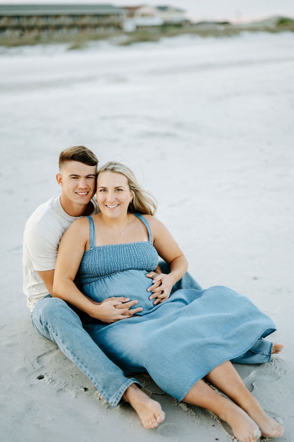 Pregnant couple sitting on beach sand, man embracing woman in blue dress holding baby bump.