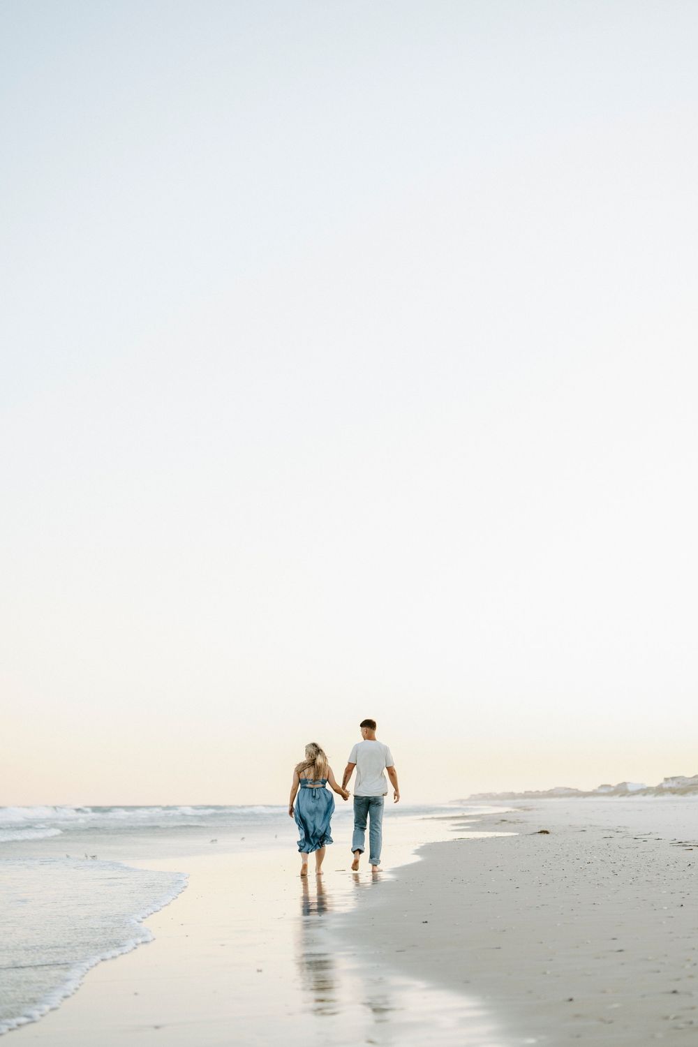 Couple holding hands walking along the shoreline at sunset on a serene beach.