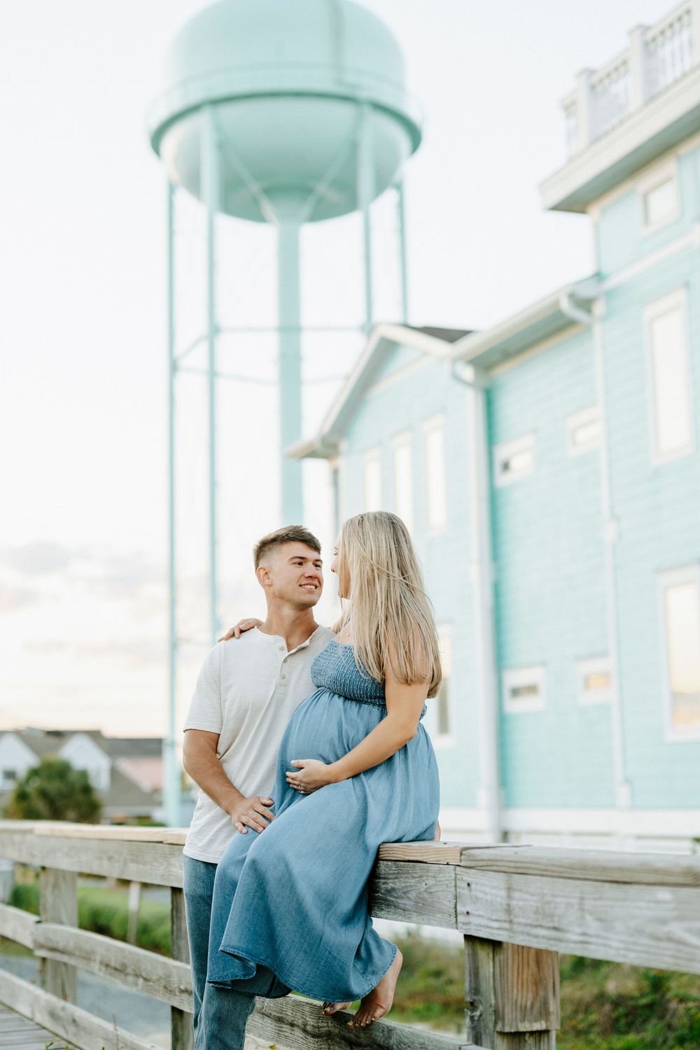 Expecting couple embracing on wooden boardwalk with teal water tower and blue building in background.