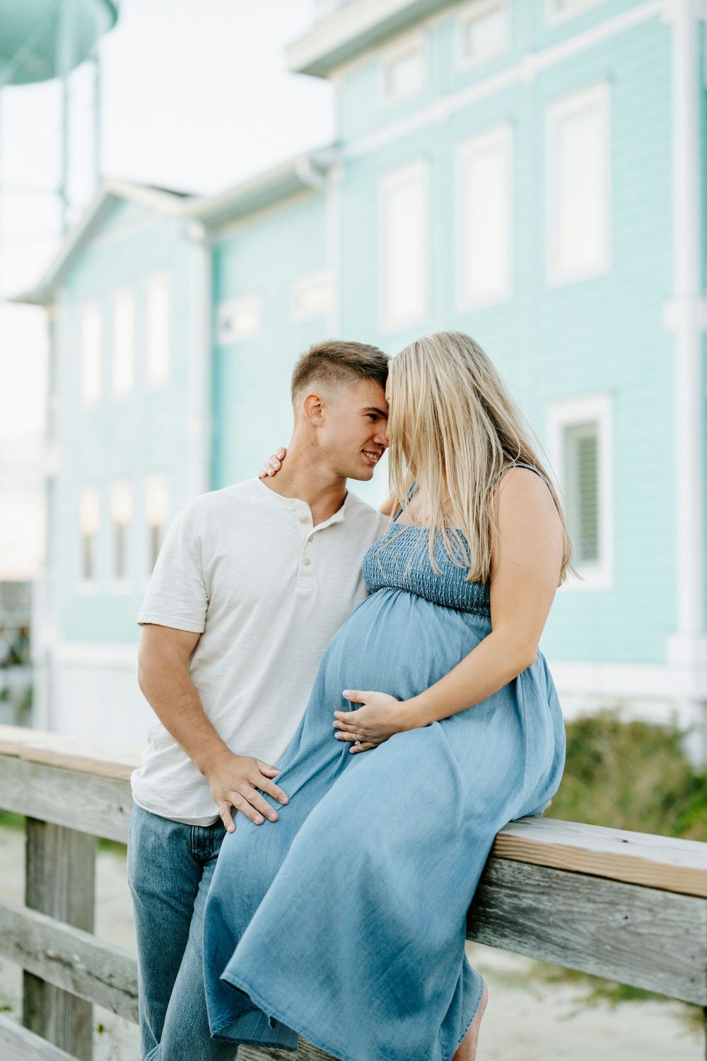 Pregnant couple sharing a tender moment on a boardwalk near a blue beach house.