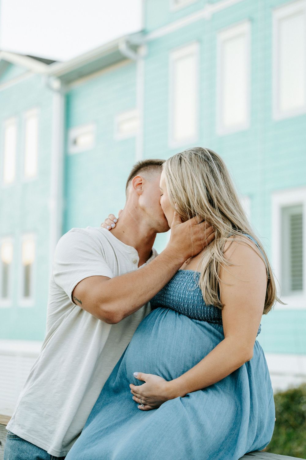 Man tenderly kissing pregnant woman in blue dress outside teal building during maternity photo session.