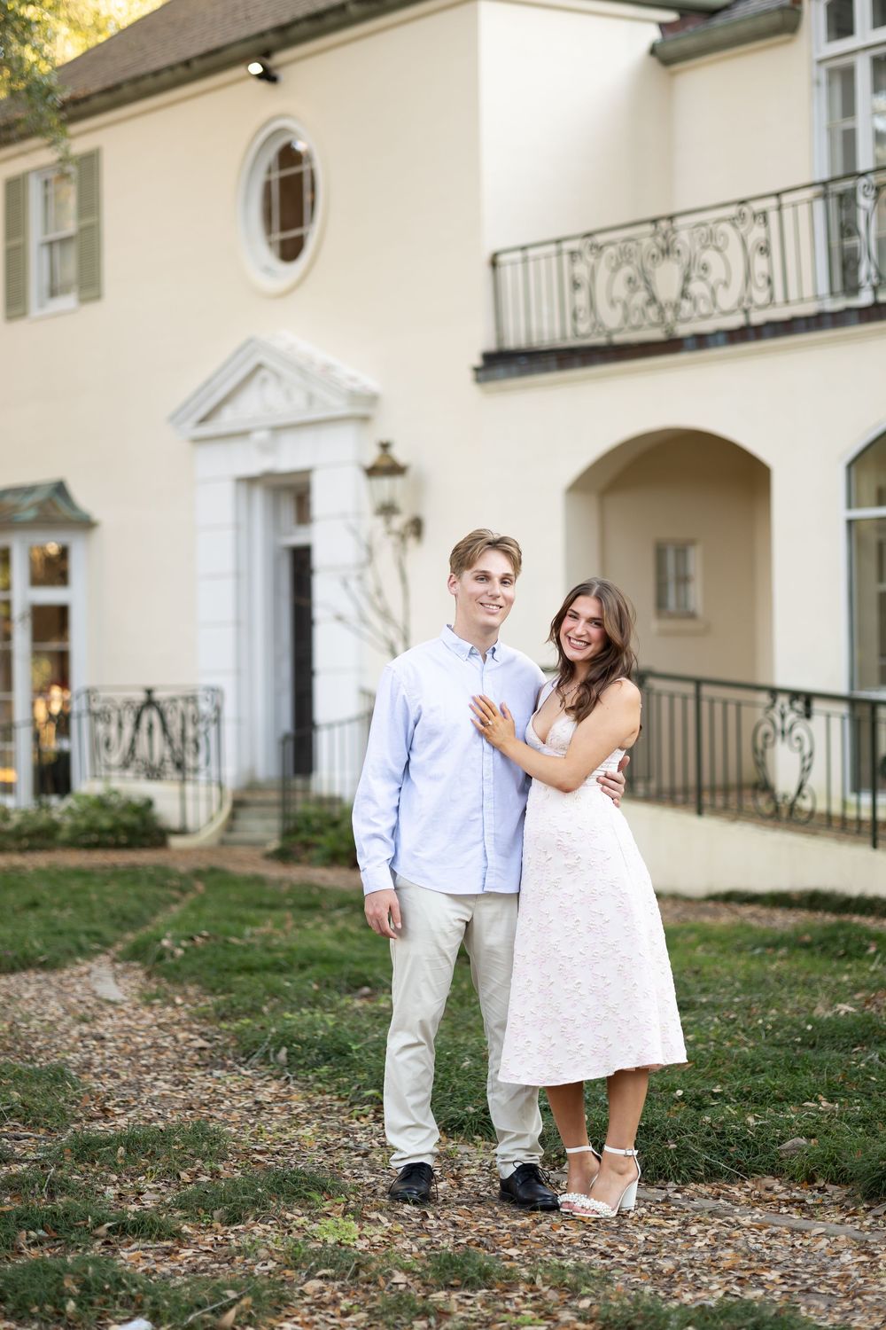 Couple posing together outside an elegant white estate, woman in white dress embracing man in light blue shirt.