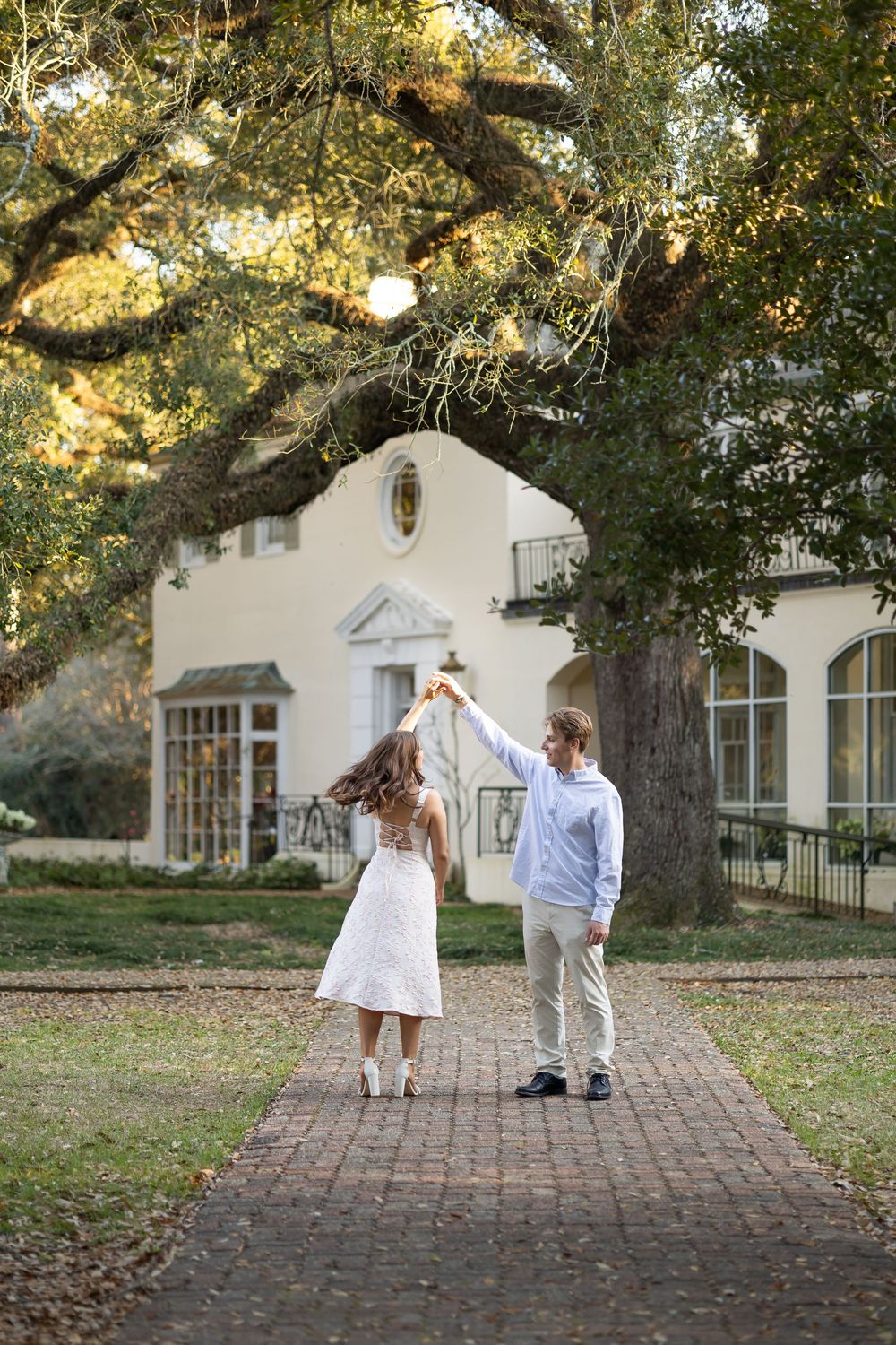 Couple dancing on brick pathway under large oak trees outside a beautiful white manor house.