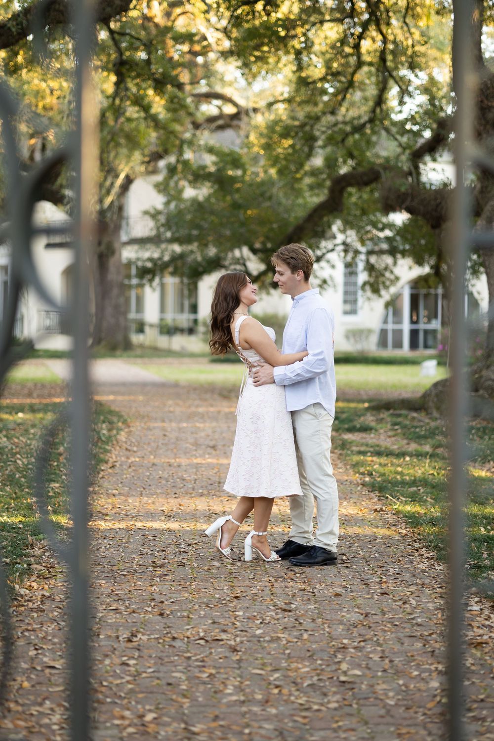 Couple embracing on a tree-lined path in a park, woman in white dress and man in light blue shirt.