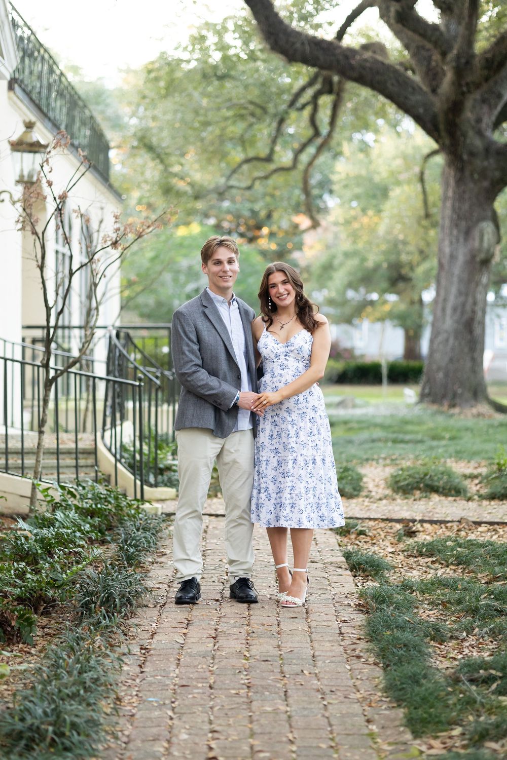 Smiling engaged couple posing on brick pathway surrounded by lush greenery, woman in floral dress, man in gray blazer.