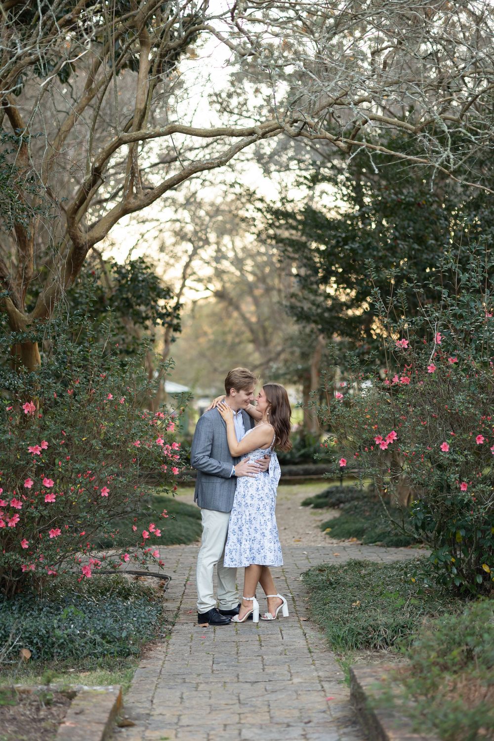 Romantic engaged couple sharing a kiss on a garden brick pathway lined with blooming pink flowers and green trees.