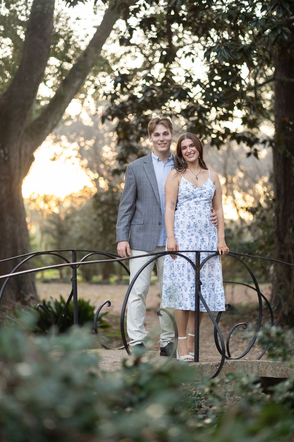 Smiling couple posing on decorative iron bridge surrounded by lush garden at sunset.