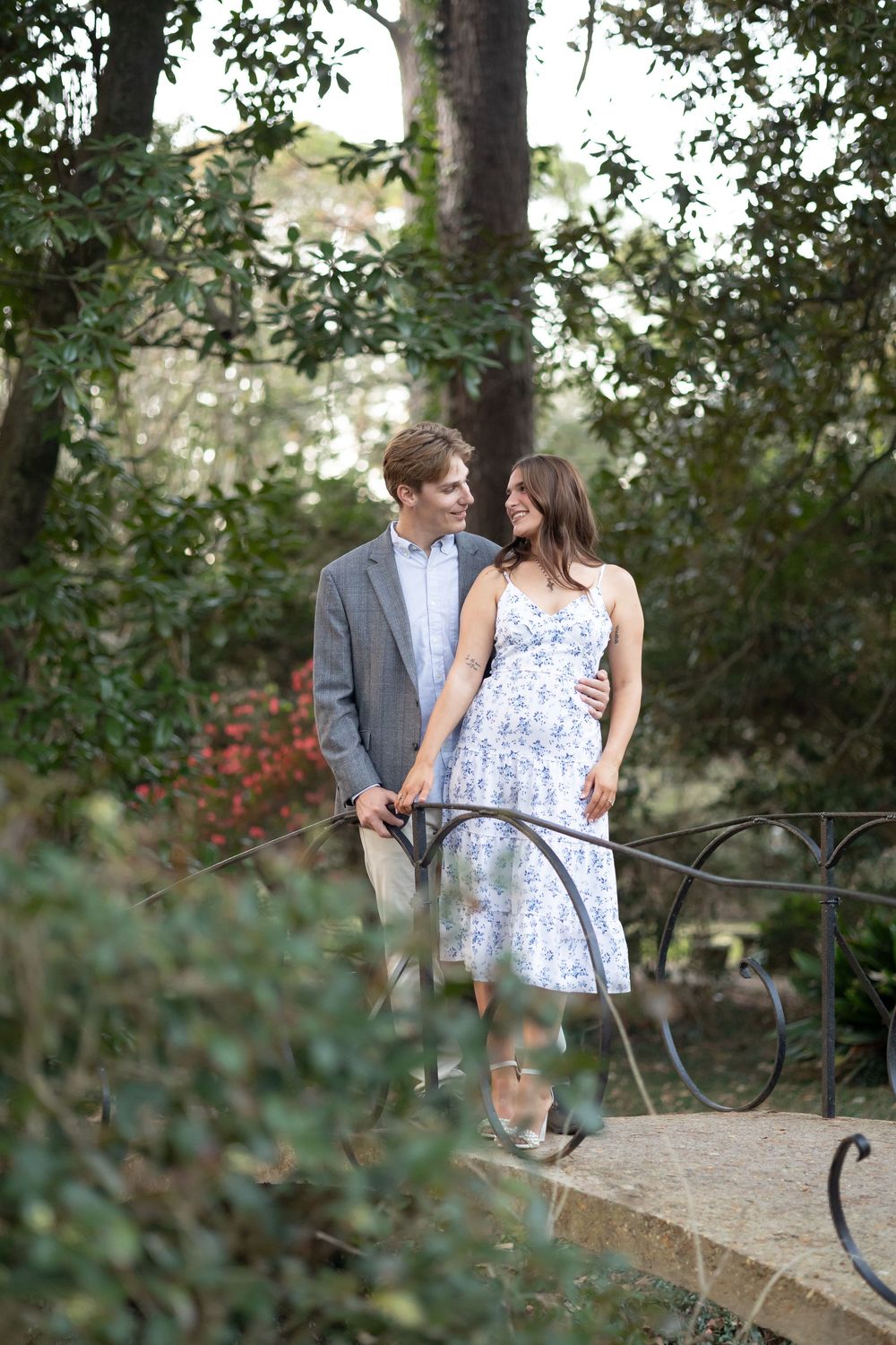 A couple shares a romantic moment on a garden bridge, surrounded by lush greenery and blooming flowers.