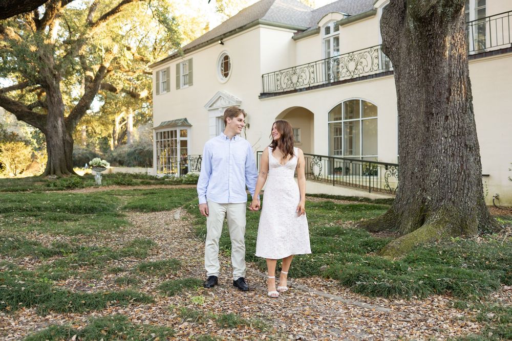 Couple holding hands and smiling while walking on a tree-lined path in front of an elegant white estate.
