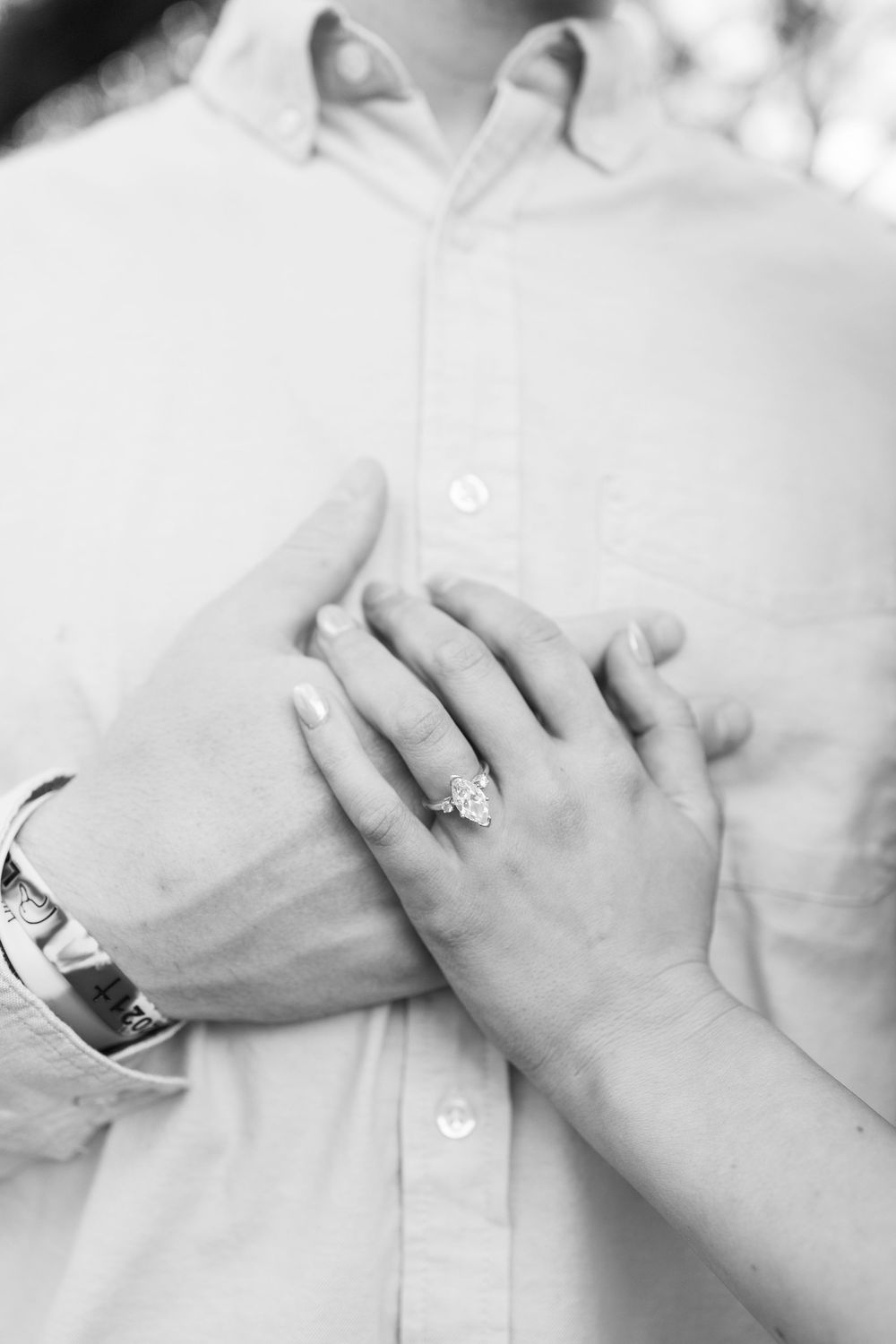 Black and white photo of engaged couple's hands showing diamond engagement ring on man's chest.