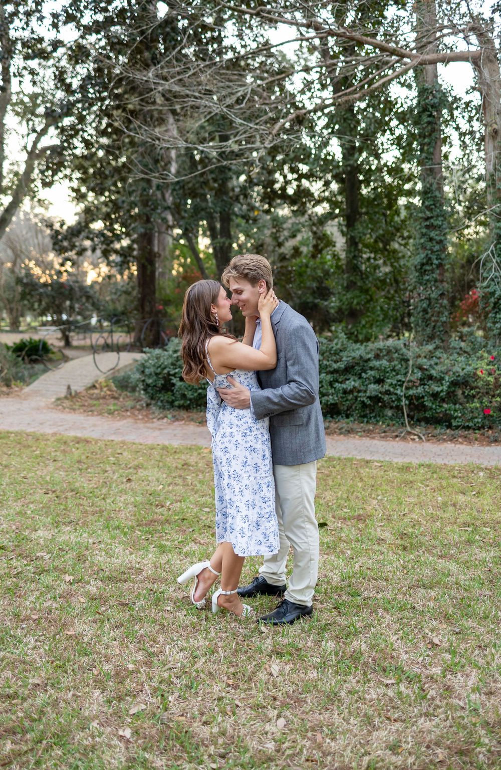 Couple embracing and sharing a romantic moment in a lush green park, woman in floral dress, man in gray blazer.
