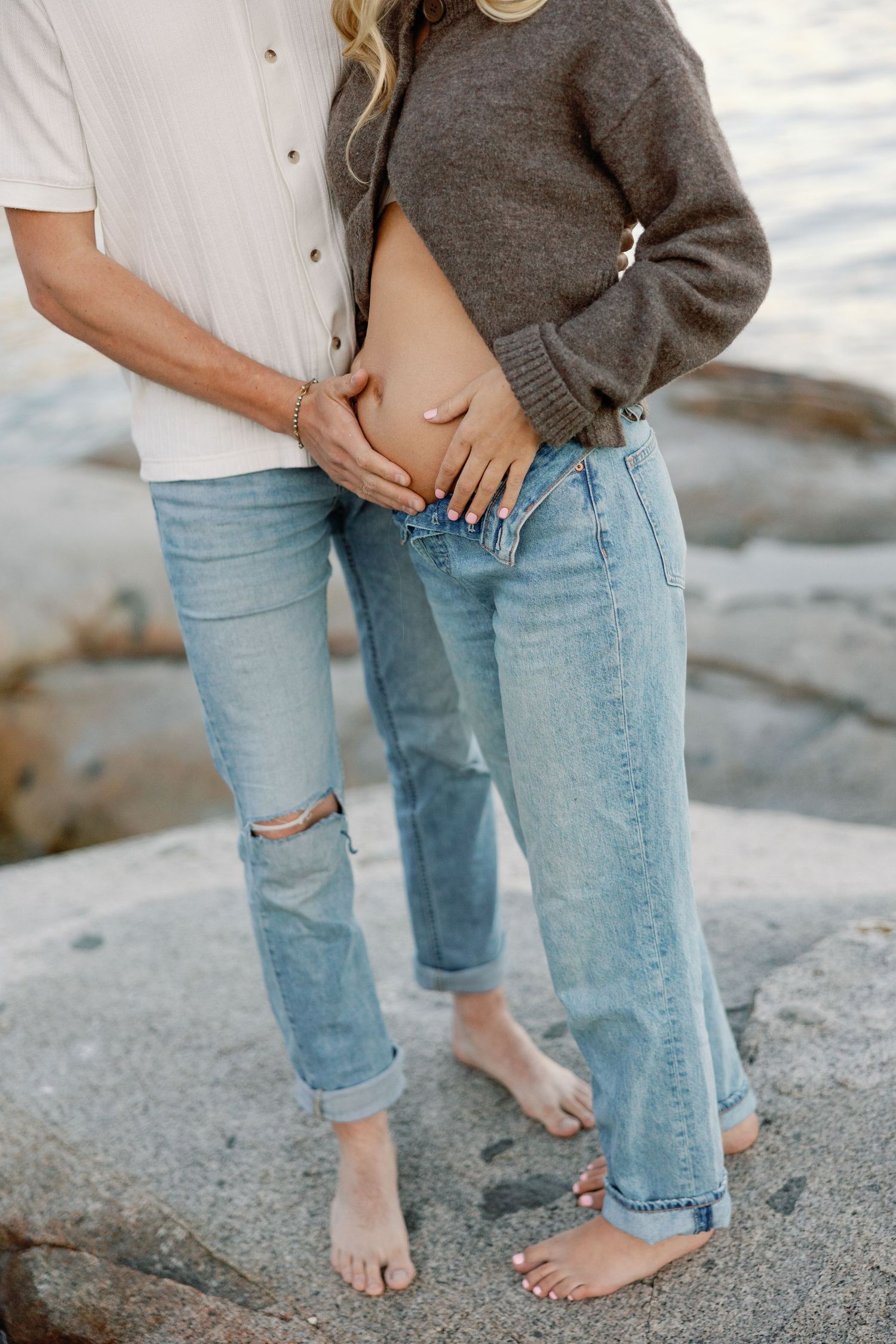 Couple standing barefoot on rocks, hands cradling pregnant belly, wearing jeans near ocean.