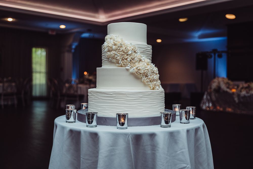 Elegant four-tier white wedding cake with floral accents on a draped table surrounded by votive candles in a reception hall.