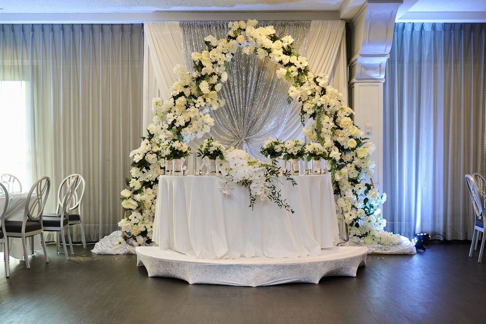 Elegant wedding sweetheart table with white floral arch, roses, and draped fabric backdrop in a reception hall.