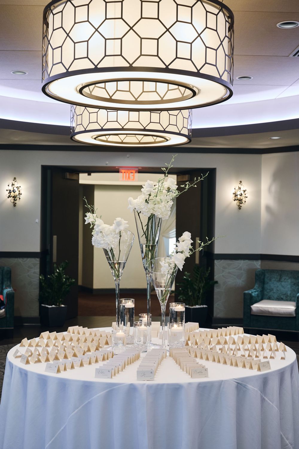 Elegant wedding escort card table with white floral centerpieces under a geometric drum chandelier in a ballroom.