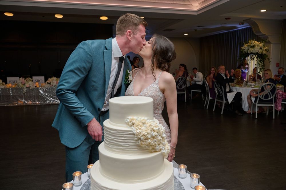 Bride and groom share a kiss behind their elegant white tiered wedding cake at their reception venue.