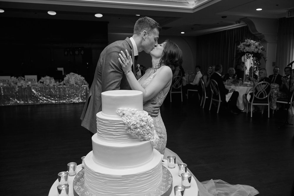 Bride and groom share a kiss behind a elegant white tiered wedding cake at their reception.