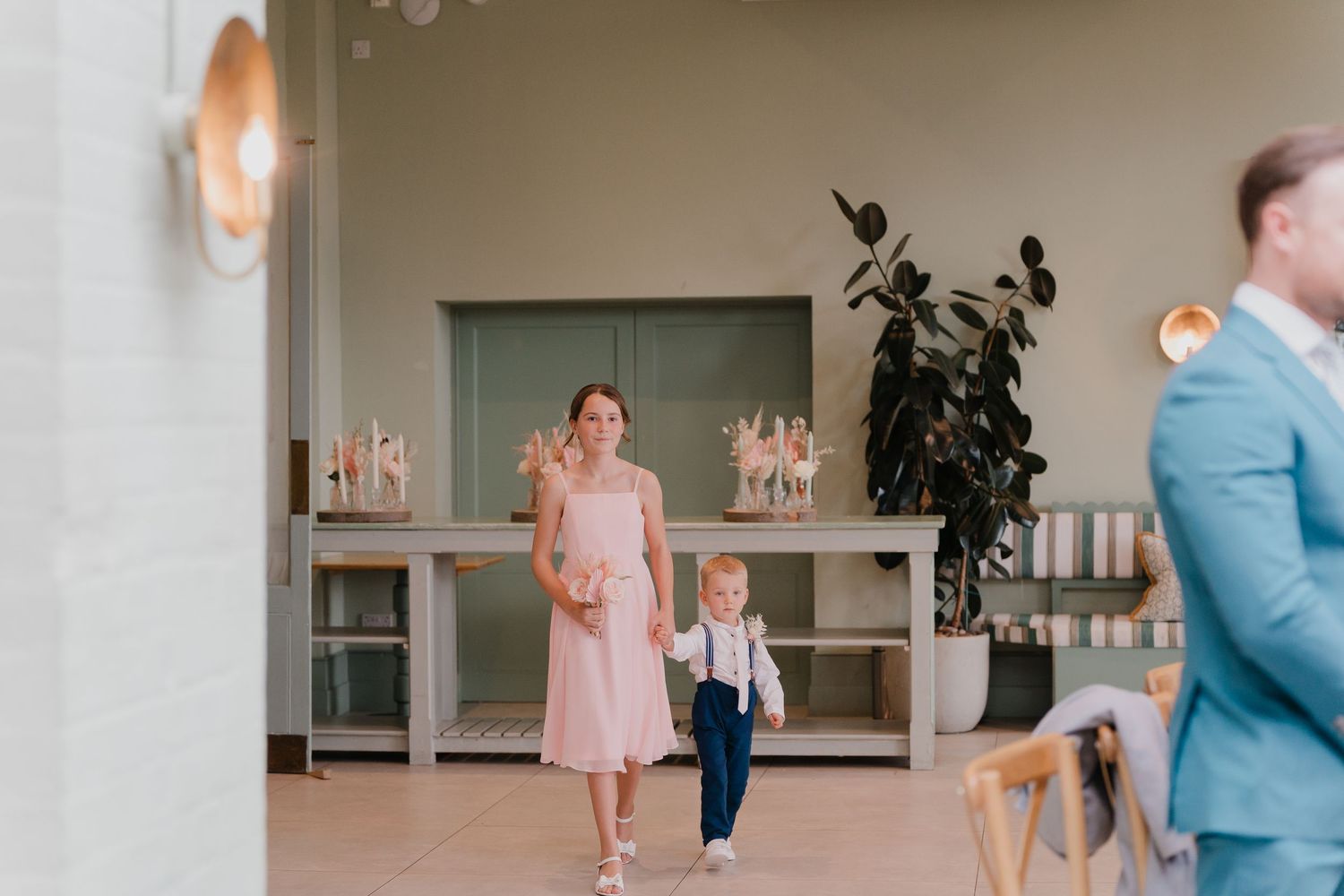 Woman in pink dress walks with young boy in suspenders through elegant venue interior.
