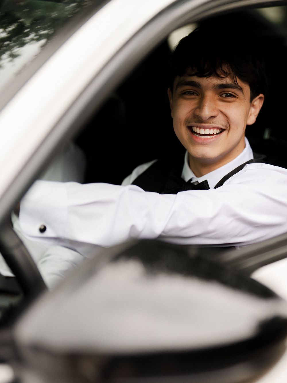 Smiling young man in white dress shirt and tie sitting in driver's seat of a car.