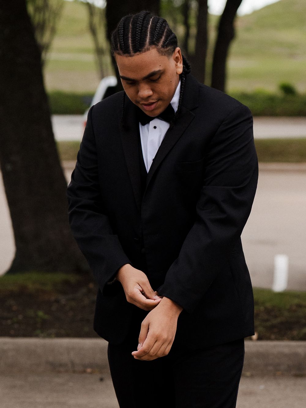 Young man in black tuxedo with bow tie adjusting his cufflinks outdoors, wearing cornrow braids.