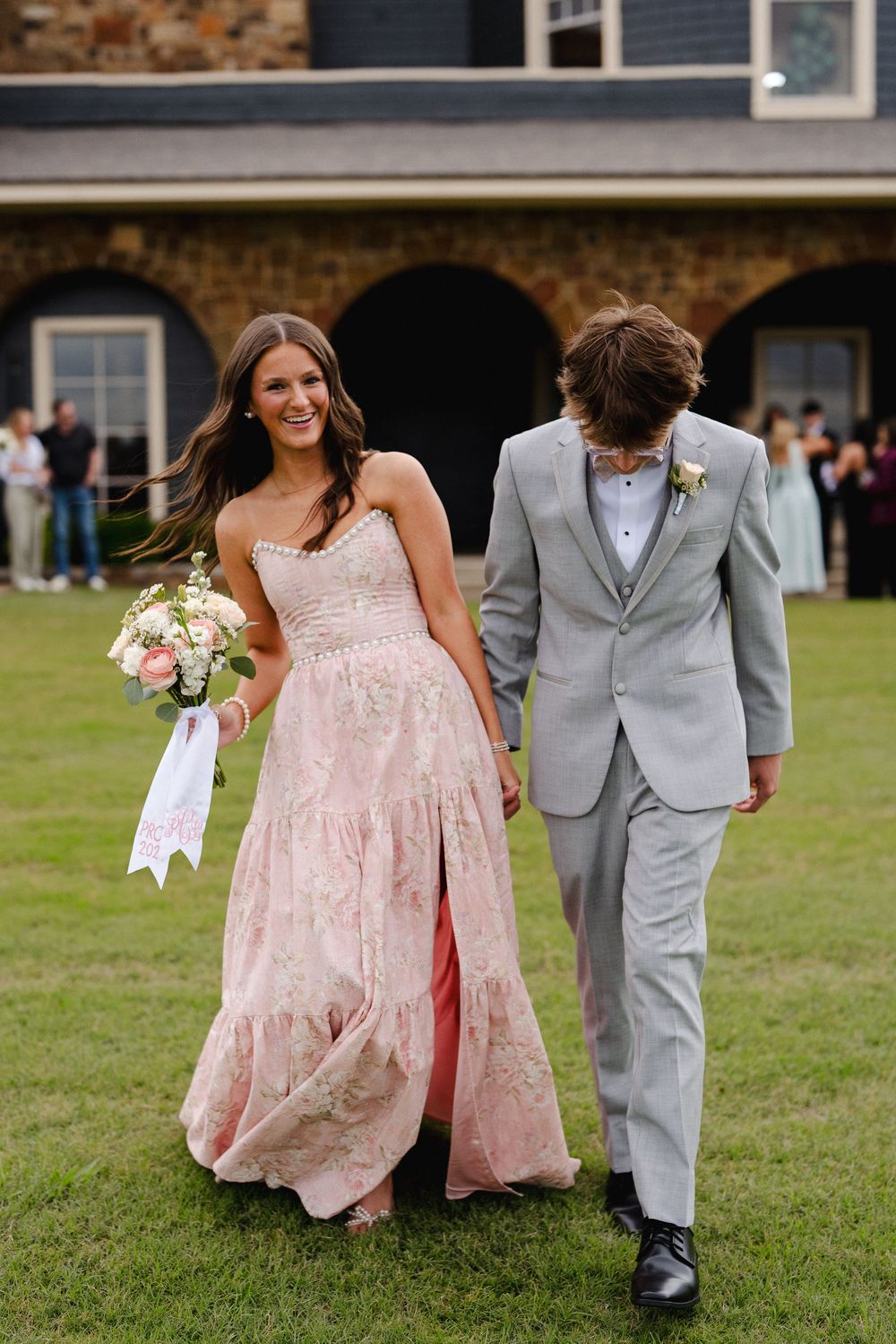 A smiling young woman in a pink floral gown holds a bouquet beside a young man in a gray suit at a prom event.