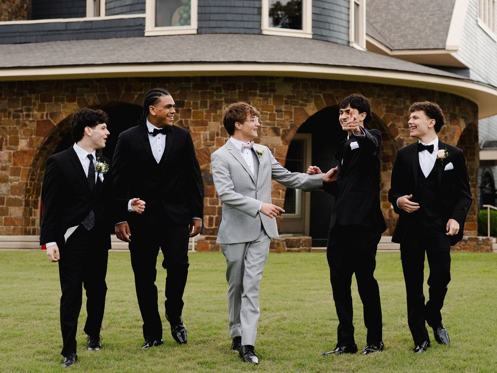 Five young men in formal tuxedos and suits pose outdoors near a stone building at a prom or wedding.