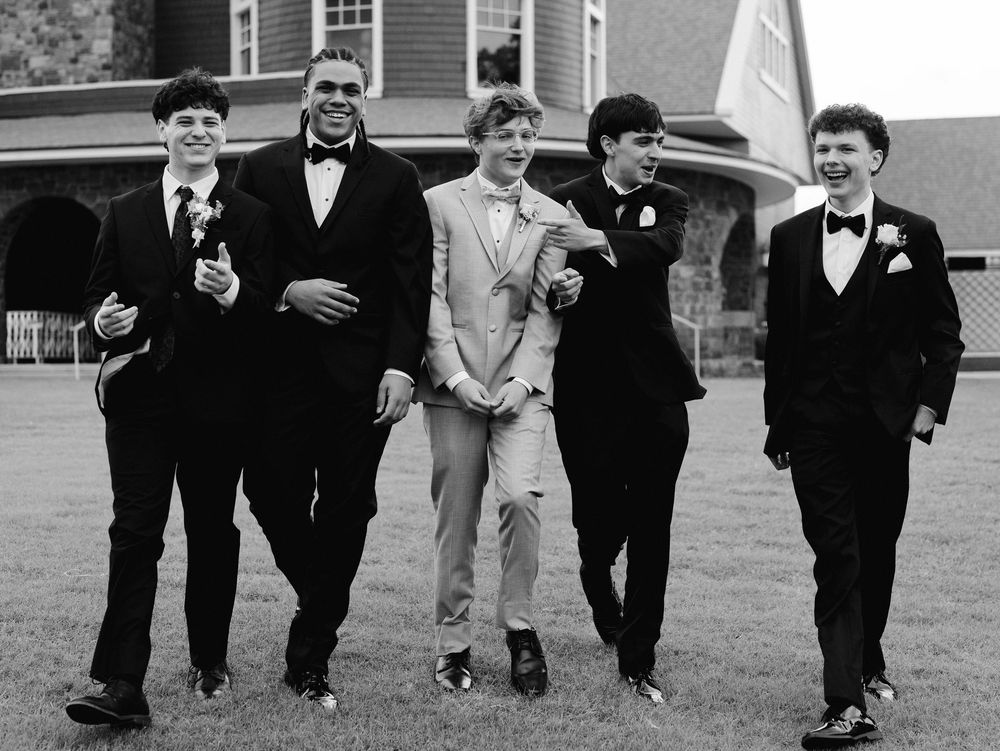 Five young men in formal attire, including tuxedos and a tan suit, walk together outdoors near a brick building.
