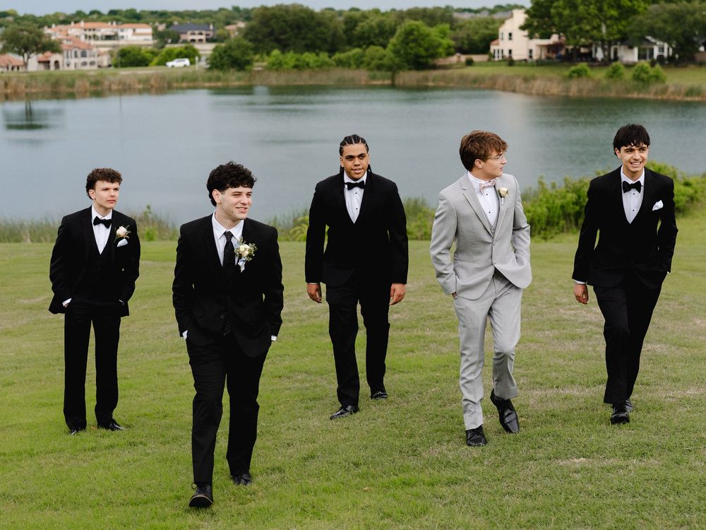Five young men in formal tuxedos walk on green grass near a lake, with one wearing a distinctive light gray suit.