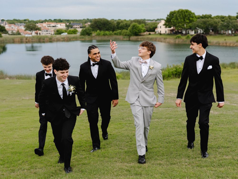 Five young men in formal tuxedos and suits walk outdoors near a lake, smiling and gesturing at a prom event.
