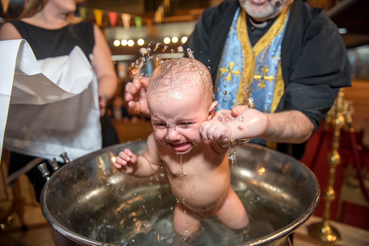 A baby sits in a large metal baptismal font during an Orthodox Christian christening ceremony.