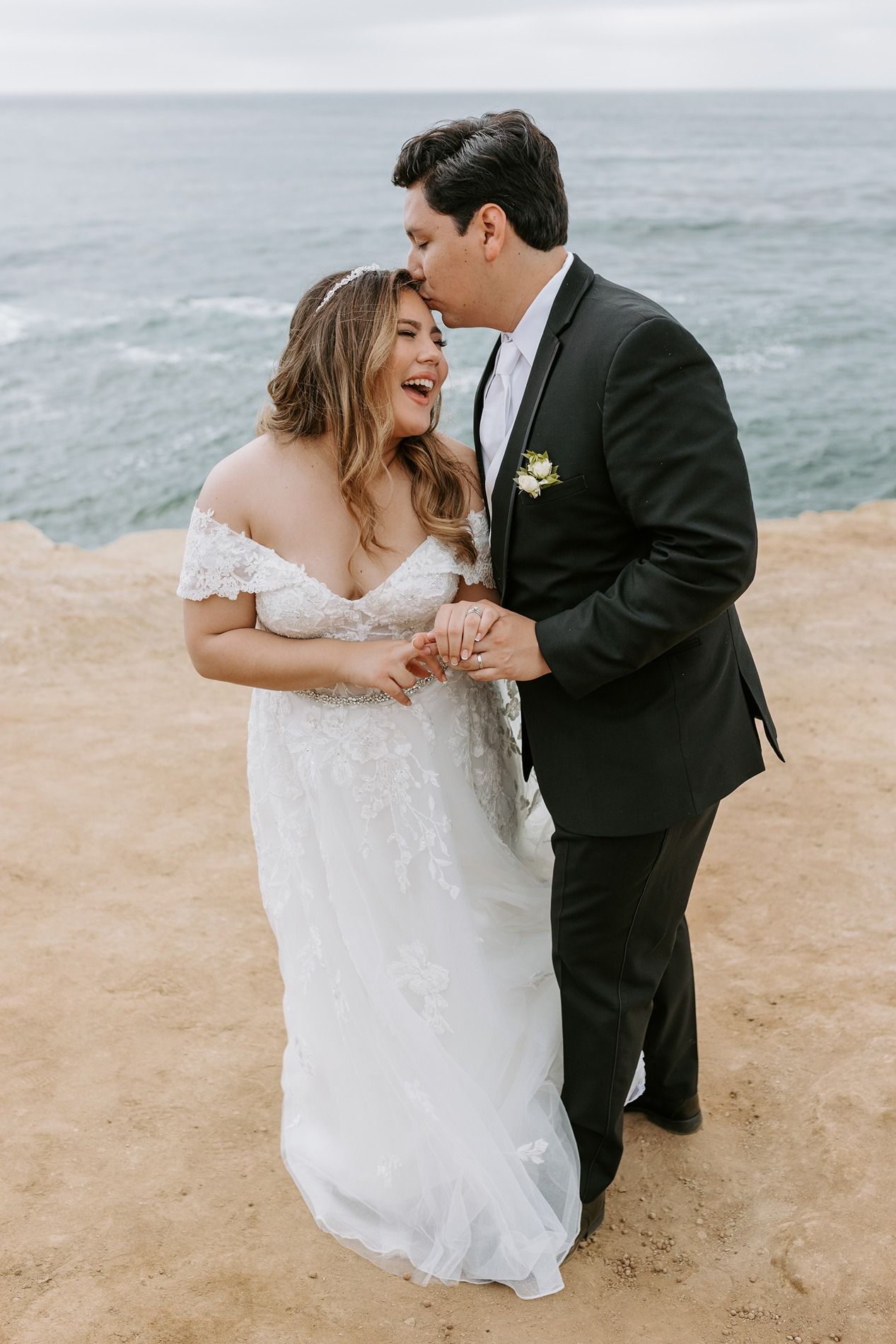 A joyful bride and groom share a romantic moment on a coastal cliff, with ocean waves in the background in San Diego.
