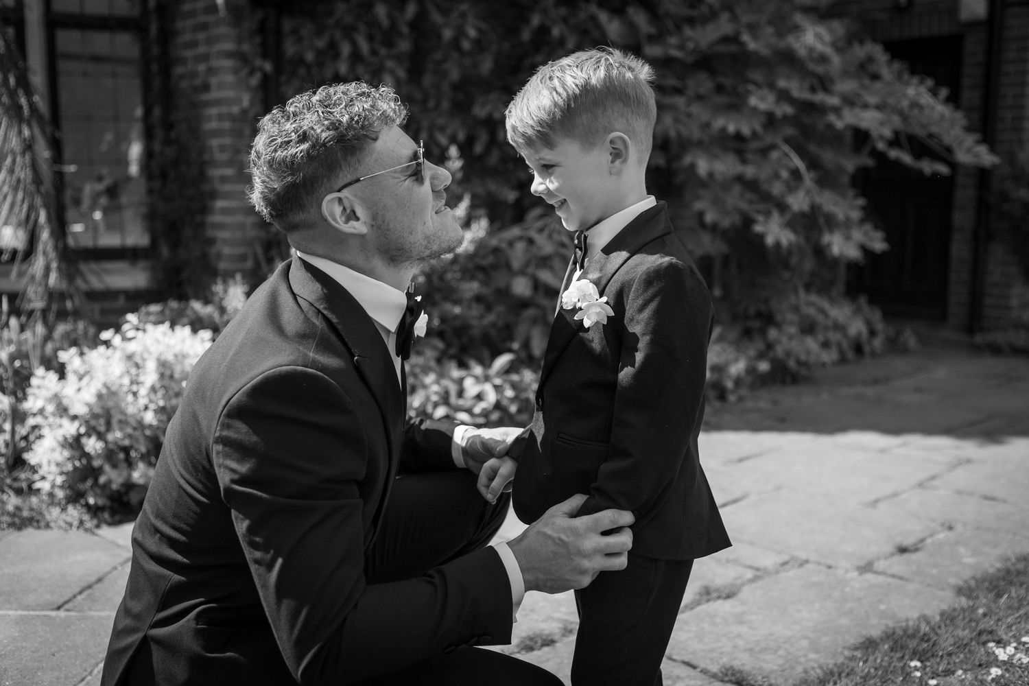 A groom in a black suit kneels down to speak with a young boy in a matching suit in a garden.