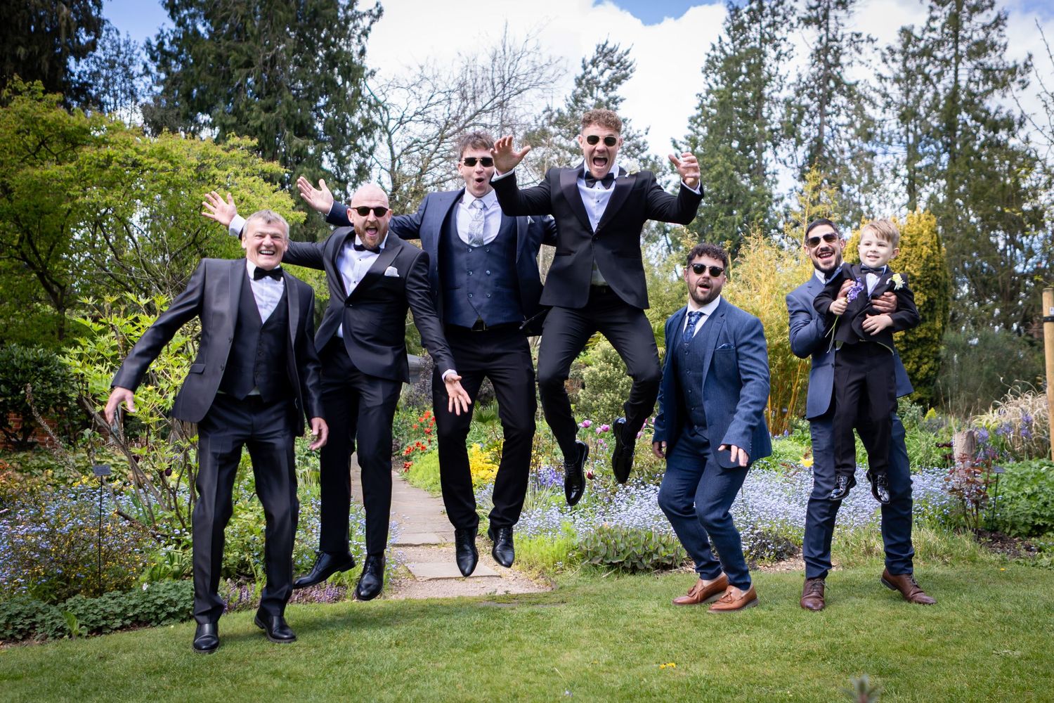 Six groomsmen in dark suits joyfully jumping in a lush garden, celebrating before a wedding ceremony.