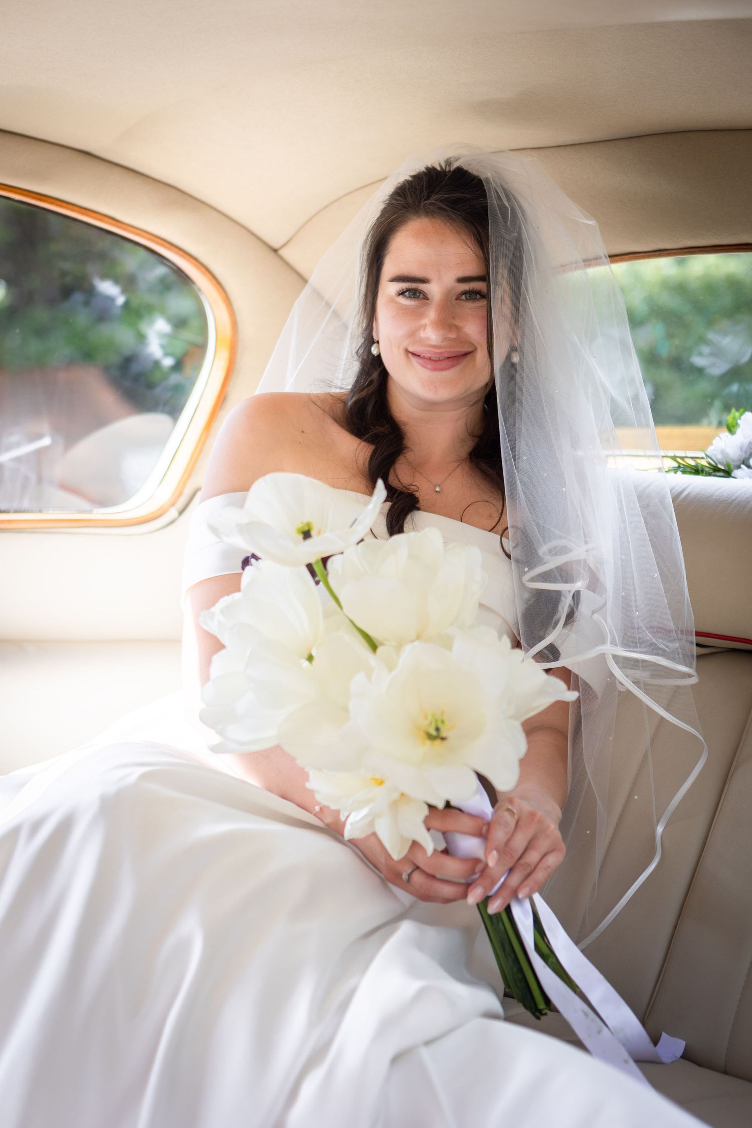 Beautiful bride in white off-shoulder gown holding white tulip bouquet while seated in a vintage wedding car.
