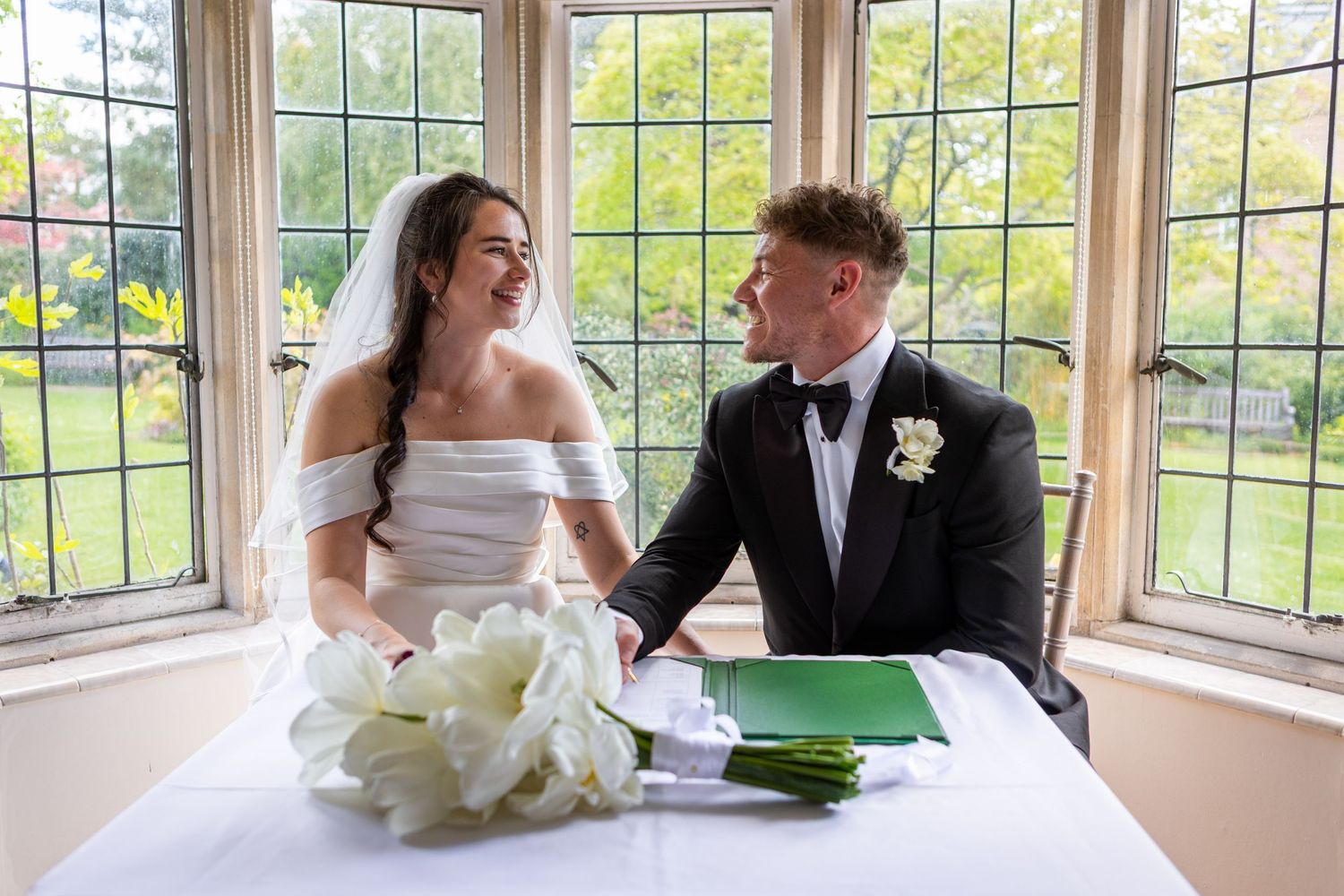 Bride and groom smiling at each other while signing the wedding register at a table by large windows.