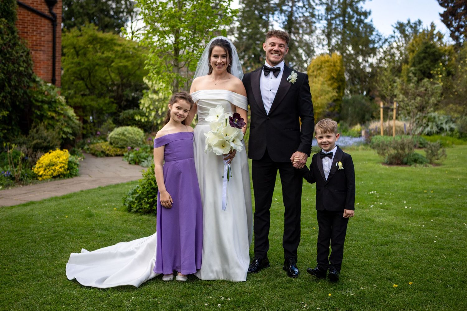 Bride and groom posing with two children in a garden, girl in purple dress and boy in black suit.
