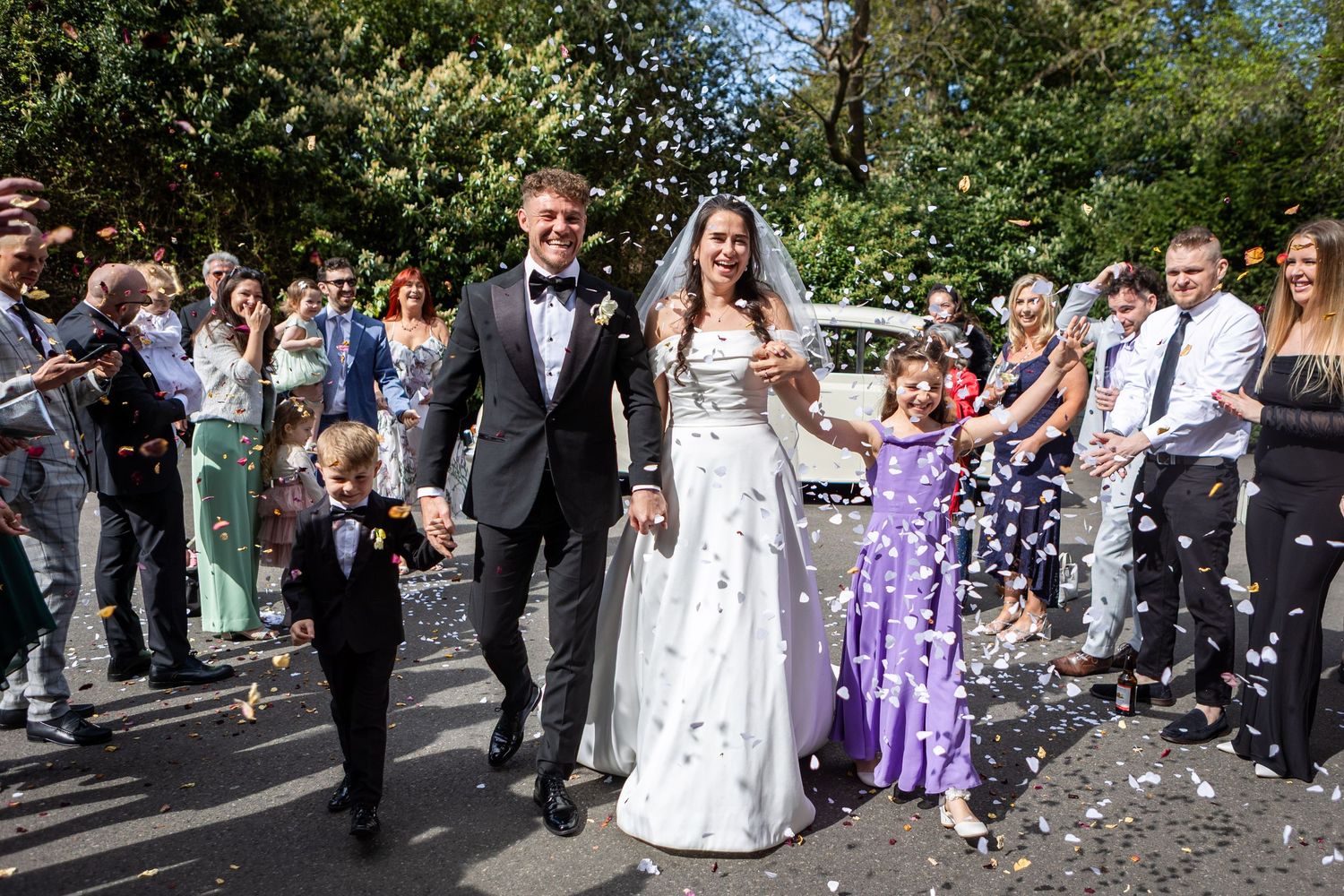Bride and groom walk joyfully through confetti shower with children, surrounded by cheering wedding guests outdoors.