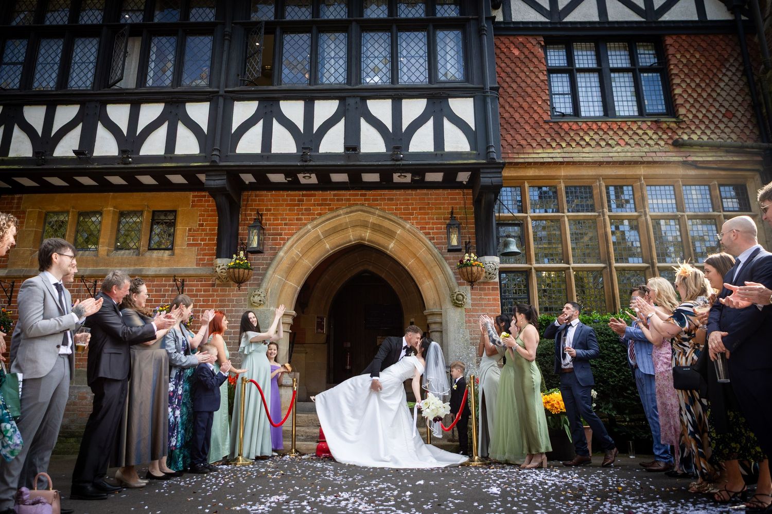 Wedding guests applaud as bride and groom exit a stunning Tudor-style venue in a shower of confetti.