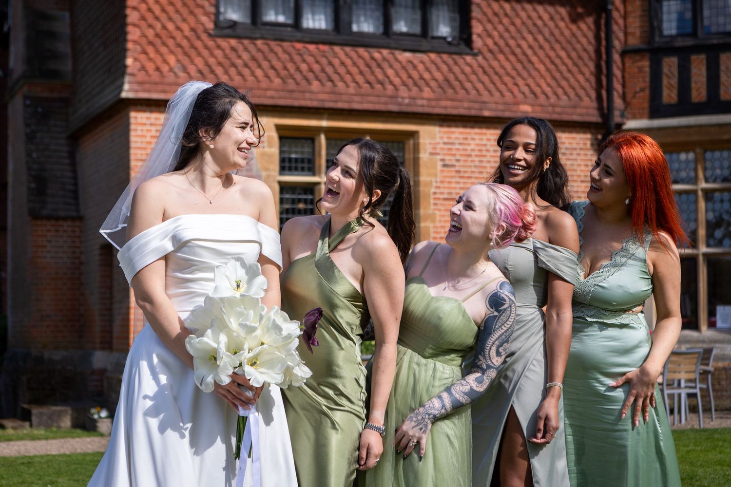 Bride in white off-shoulder gown holds bouquet while laughing with four bridesmaids in sage green dresses outside brick manor.