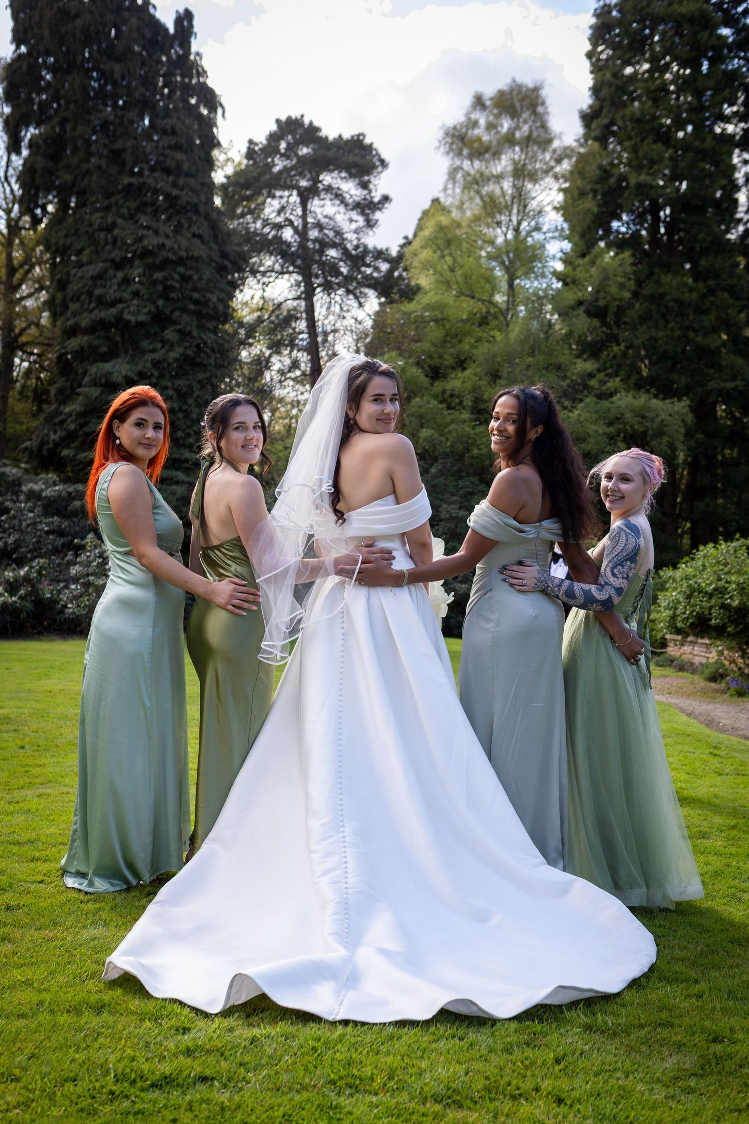 Bride in white gown and veil poses with four bridesmaids in sage green dresses in a lush garden setting.