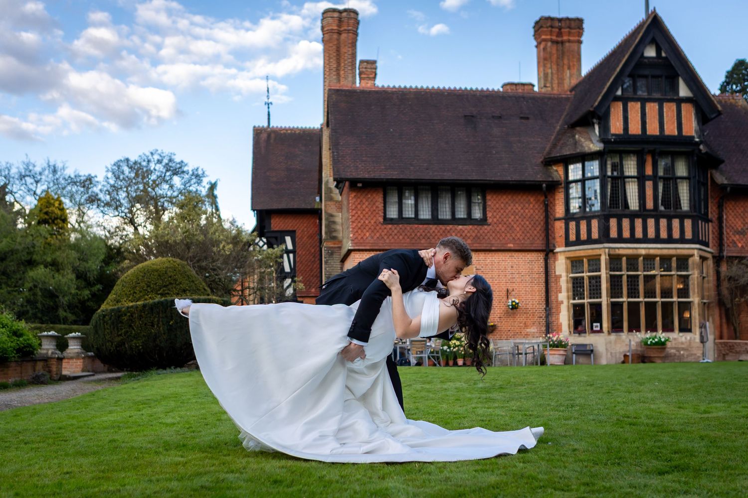 Groom dips bride in white dress for romantic photo outside a Tudor-style manor house at golden hour.