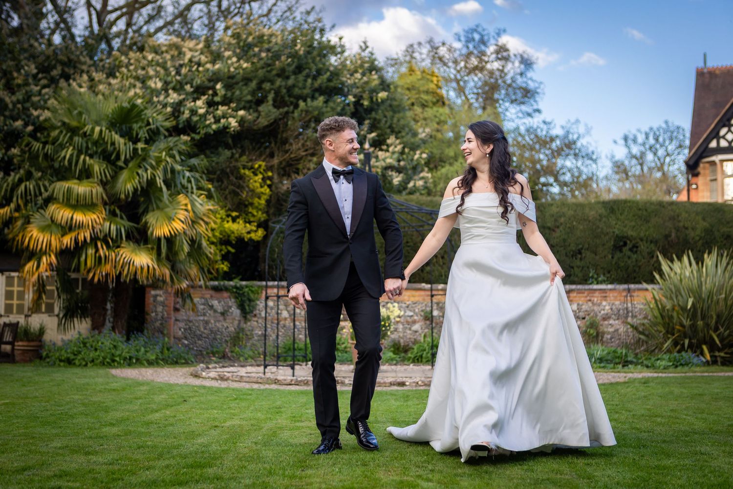Bride and groom hold hands walking through garden, she in white off-shoulder gown, he in black tuxedo.