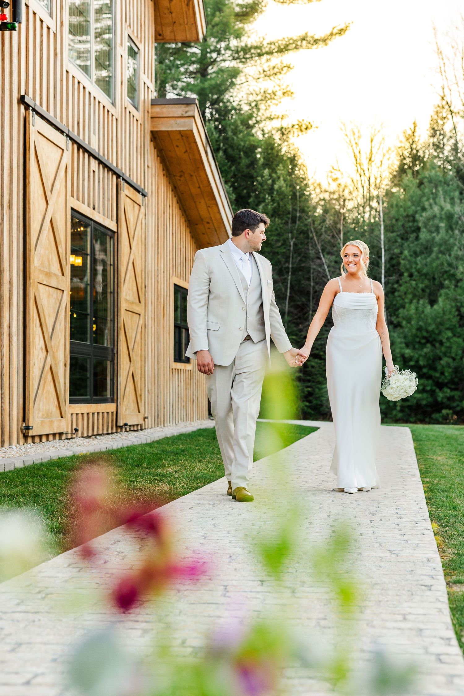 A bride and groom walk hand-in-hand along a stone path beside a wooden barn venue at golden hour.