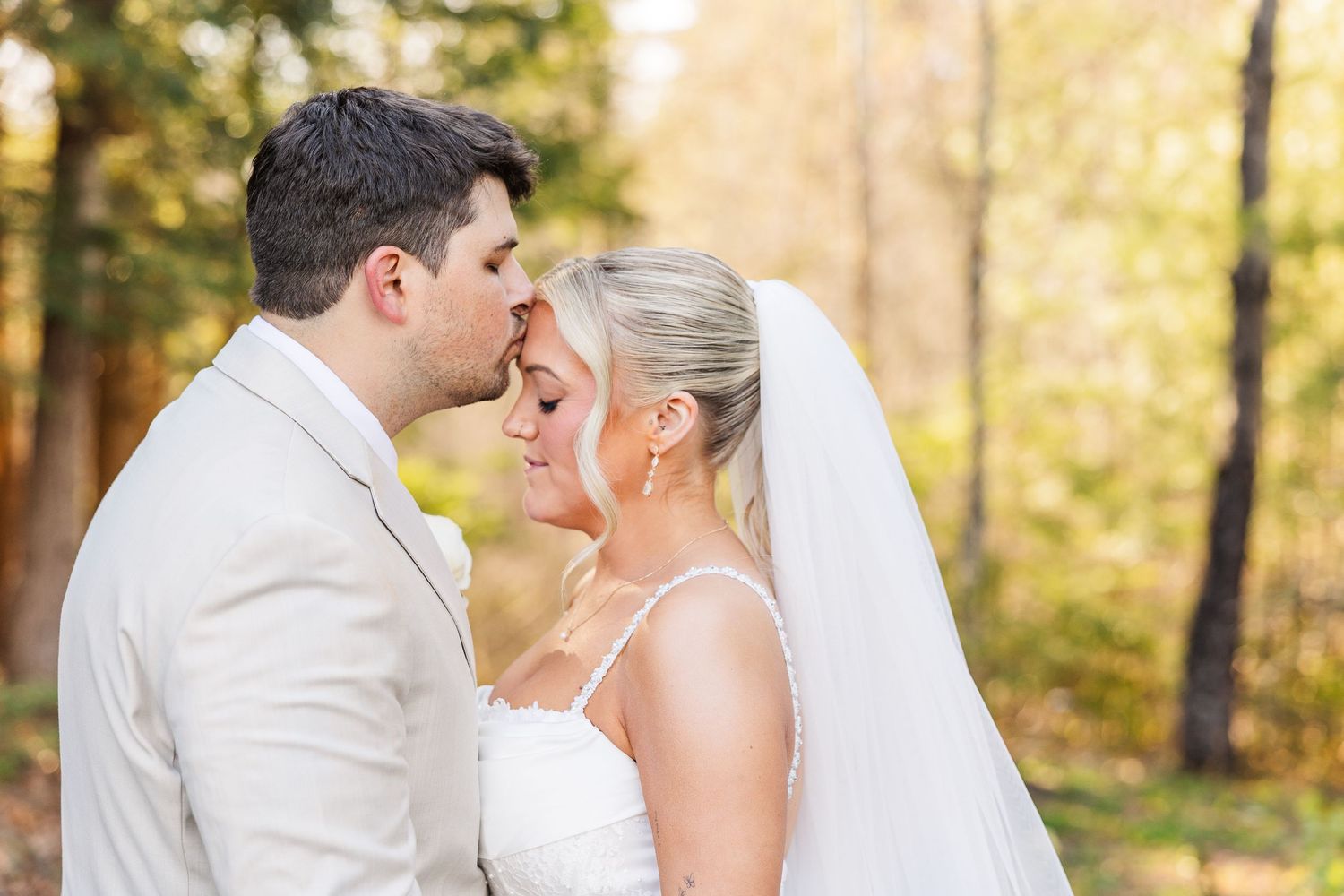 Groom in white suit kissing bride on forehead outdoors, bride wearing veil and strapless gown with autumn trees.