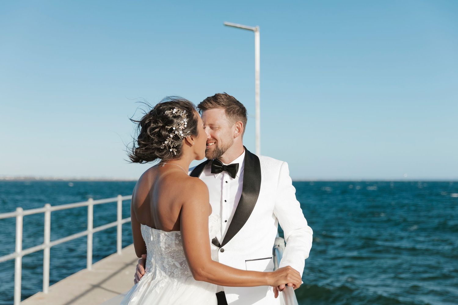 Bride and groom share a romantic kiss on a pier overlooking the ocean, groom wearing a white tuxedo jacket.