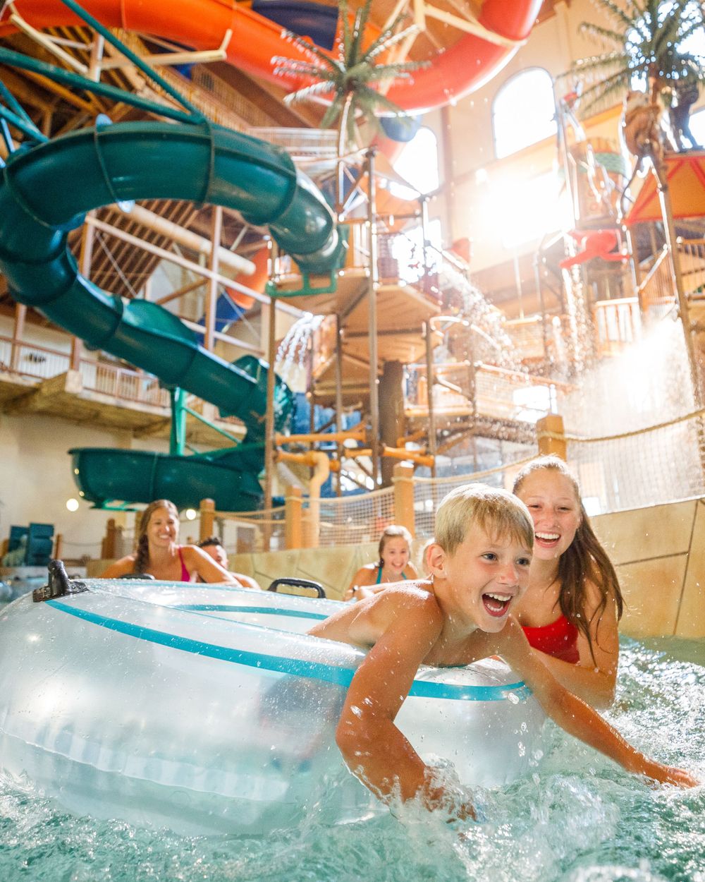 Children having fun splashing and riding tubes at an indoor water park with colorful waterslides and bright sunlight streaming in.