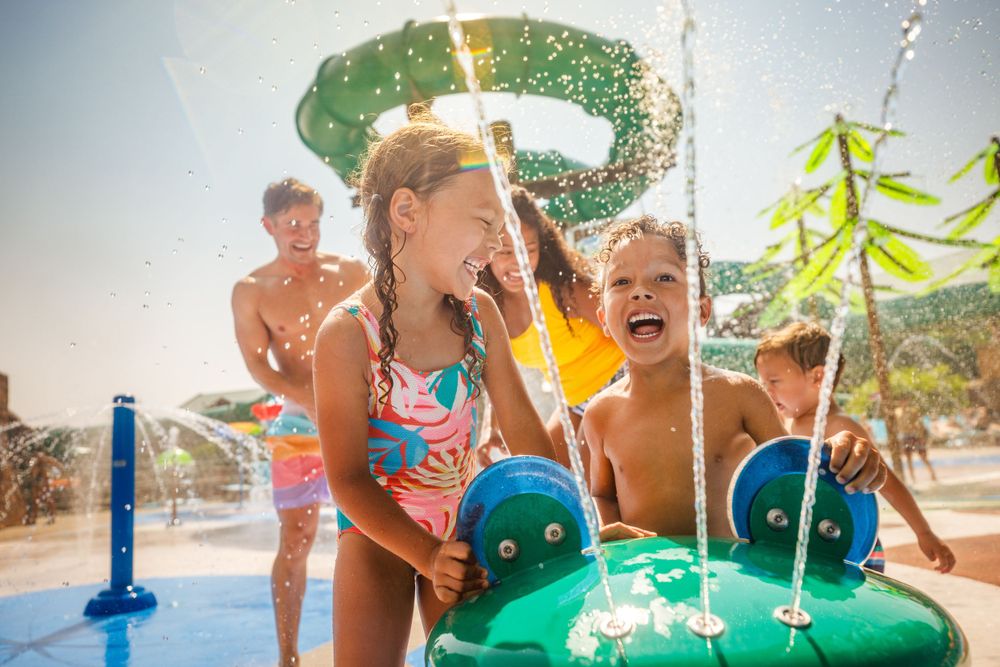 Children playing and splashing at a water park on a bright sunny summer day with water features and fountains.