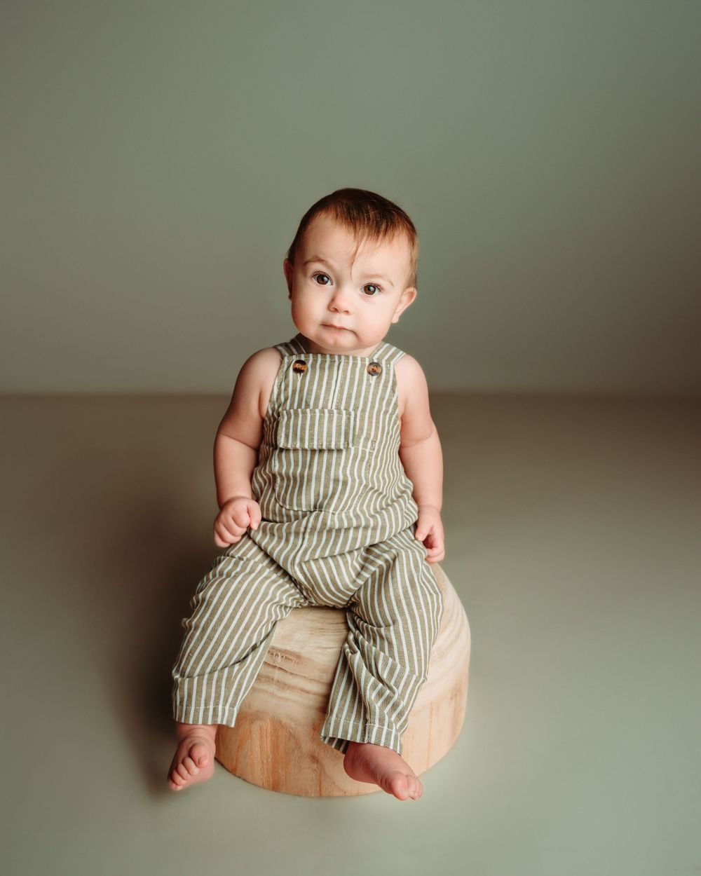 Studio portrait in striped linen overalls sitting on wooden stool.